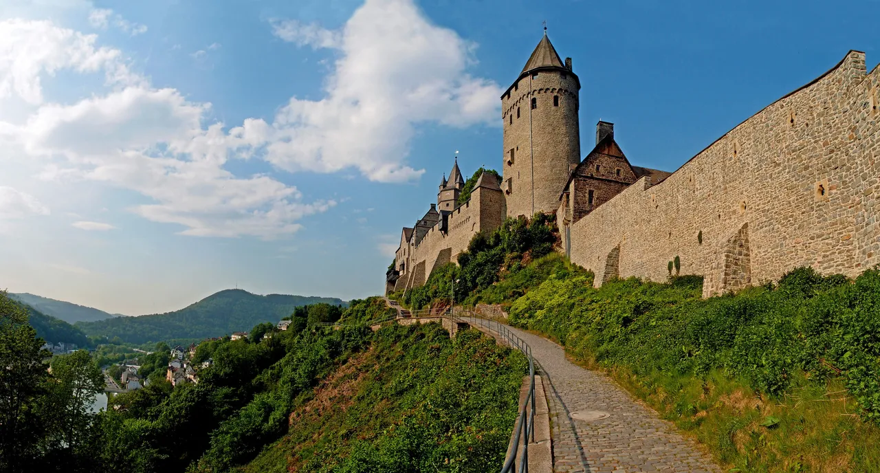 Die Burg Altena erhebt sich auf einem Hügel über einer Stadt und Landschaft, ein steinernes Gebäude mit Türmen und Mauern unter einem blauen Himmel mit Wolken.