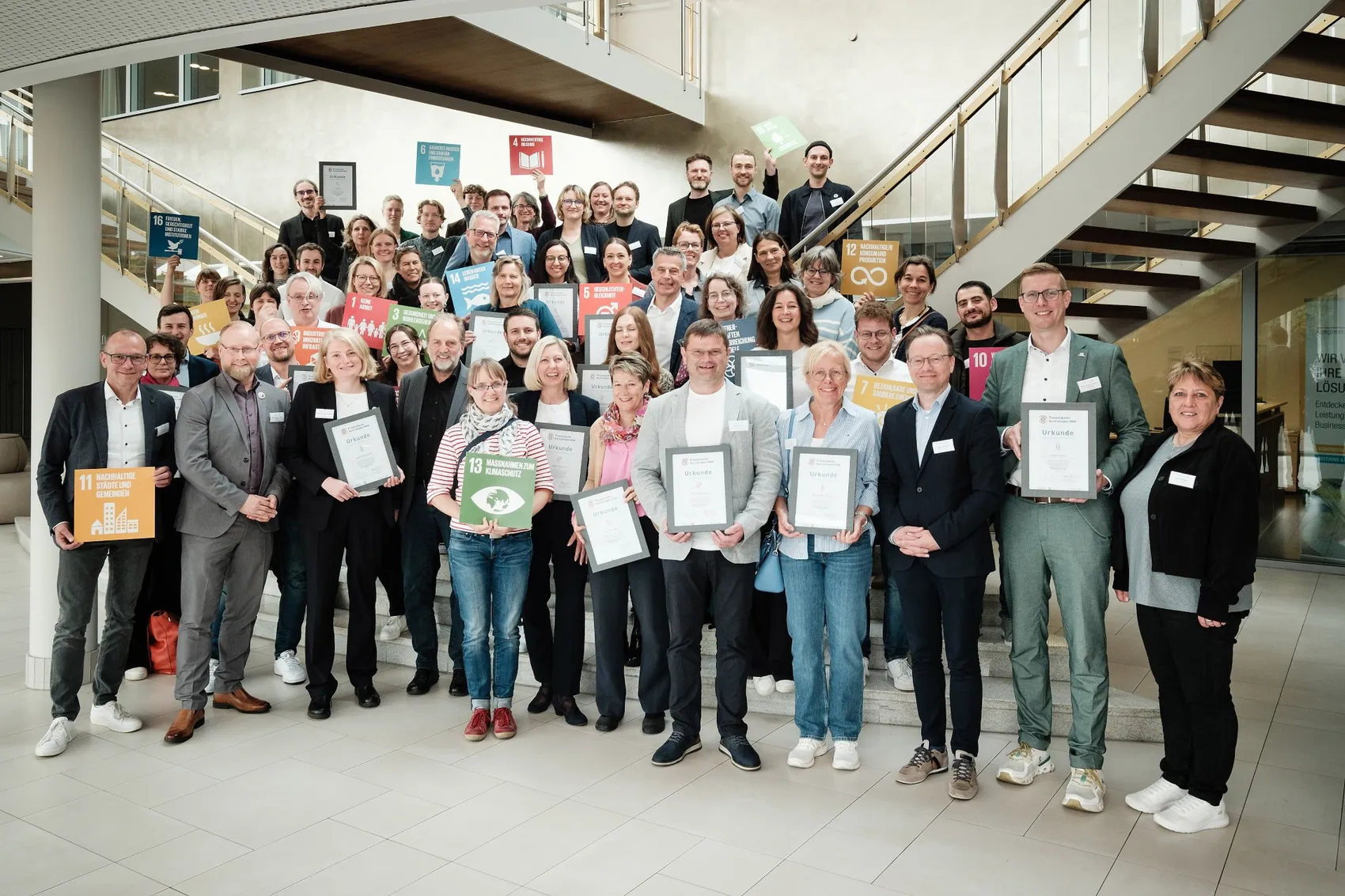 Eine Gruppe von etwa 40 Personen steht auf mehreren Treppenstufen in einem hellen Innenraum zusammen und hält Urkunden in den Händen. Im Hintergrund sind Banner mit Symbolen für Nachhaltigkeitsziele zu sehen.