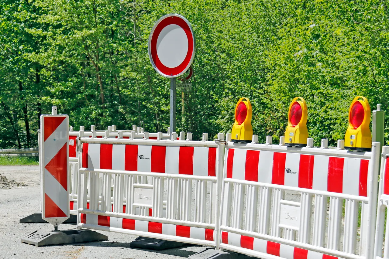 Rot-weiße Absperrgitter mit gelben Warnleuchten und ein rundes Verkehrszeichen „Verbot der Einfahrt“ vor grünen Bäumen.