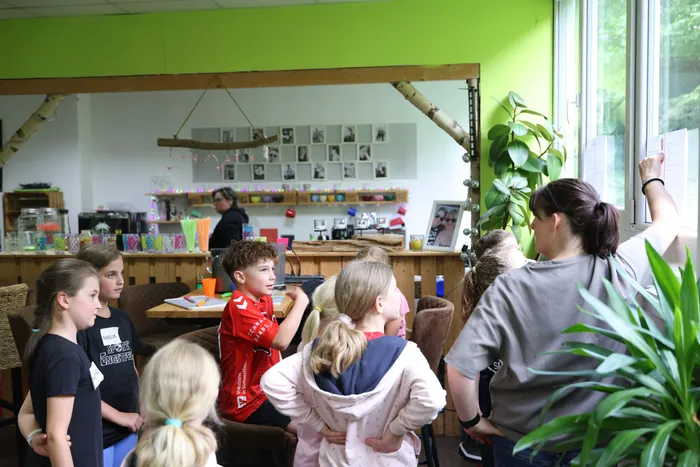 Eine Frau zeigt Kindern in einem hellen Raum ein Blatt Papier mit Informationen. Die Kinder stehen in einer Gruppe und schauen auf das Papier, während im Hintergrund weitere Kinder und Erwachsene sitzen.