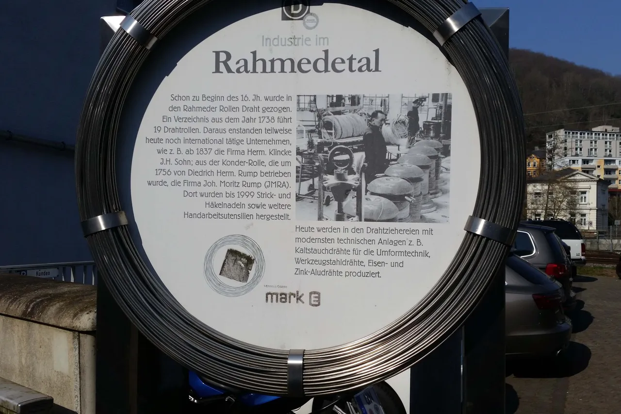 Eine Informationstafel mit dem Titel „Industrie im Rahmedetal“ zeigt historische und moderne Bilder der Drahtproduktion. Im Hintergrund ist die Talsperre Fuelbecke zu sehen.