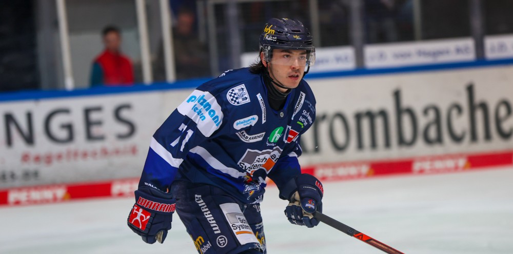 Eishockeyspieler Daniel Fischbuch in voller Ausrüstung mit Schläger auf dem Eis, während eines Spiels. Im Hintergrund sind Zuschauer und Werbebanden zu sehen.