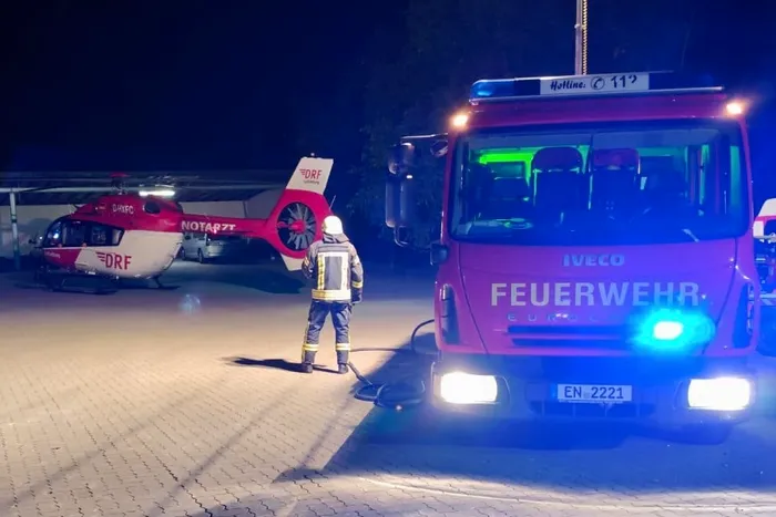 Ein Feuerwehrmann steht nachts neben einem Feuerwehrauto vor einem DRF-Rettungshubschrauber auf einem Landeplatz.
