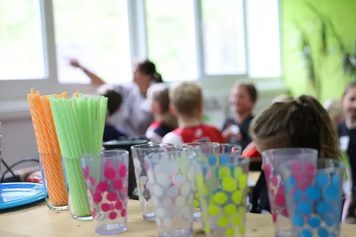 Bunte Trinkgläser und Strohhalme stehen im Vordergrund, während im Hintergrund Kinder und eine Betreuerin an einem Workshop teilnehmen.