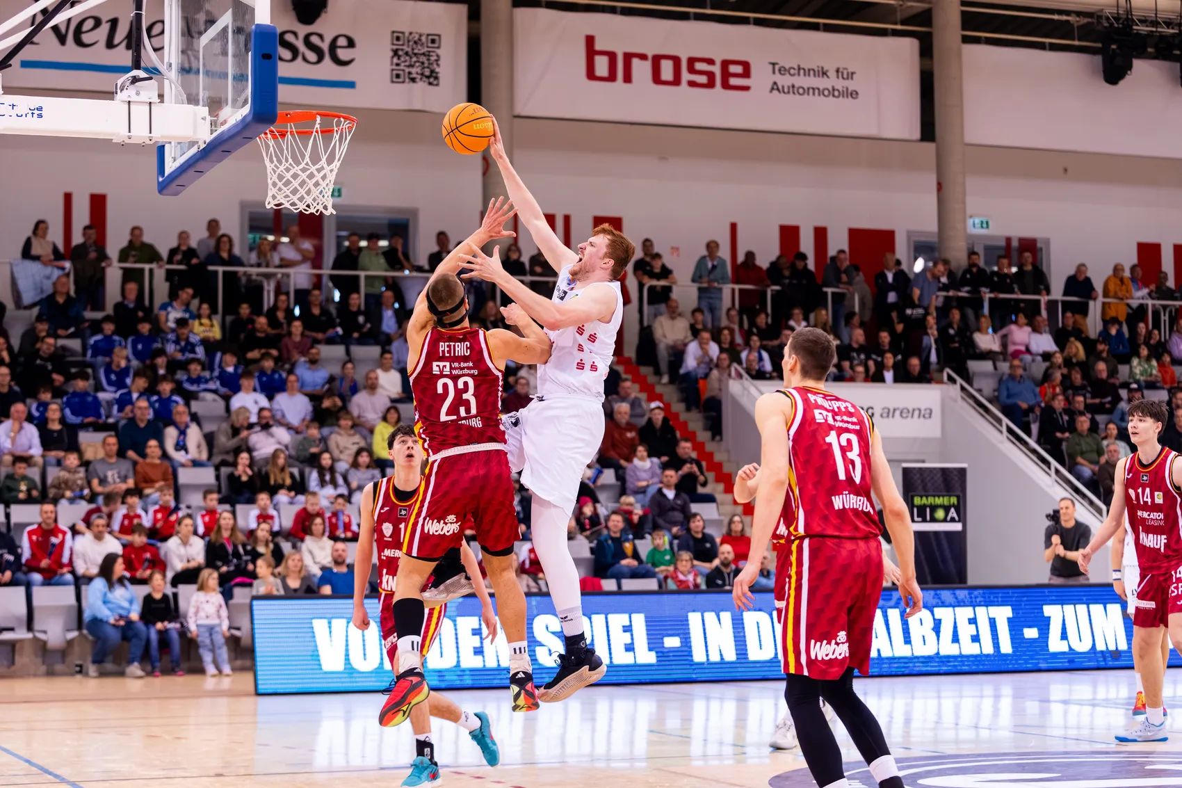 Ein Basketballspieler in einem weißen Trikot wirft den Ball während eines Spiels in einer vollen Halle. Mehrere Gegenspieler in roten Trikots versuchen, den Wurf zu blocken. Im Hintergrund ist ein Banner mit der Aufschrift „VON DER SPIEL-IN DER ALBZEIT - ZUM“ zu sehen.