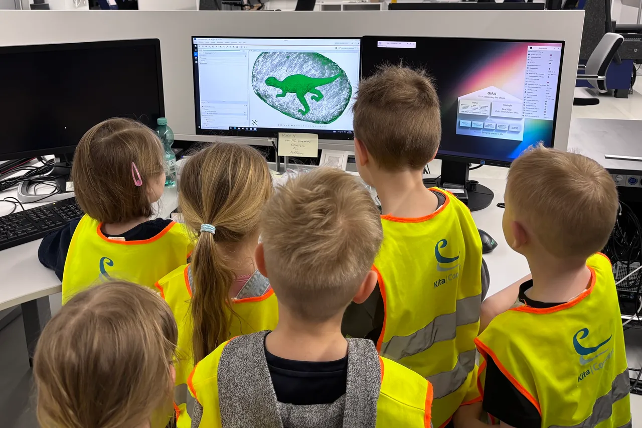 Eine Gruppe von sechs Kindern mit gelben Westen steht vor einem Computerbildschirm und betrachtet ein Bild eines Dinosauriers. Im Hintergrund sind weitere Computer und Büromaterialien zu sehen.