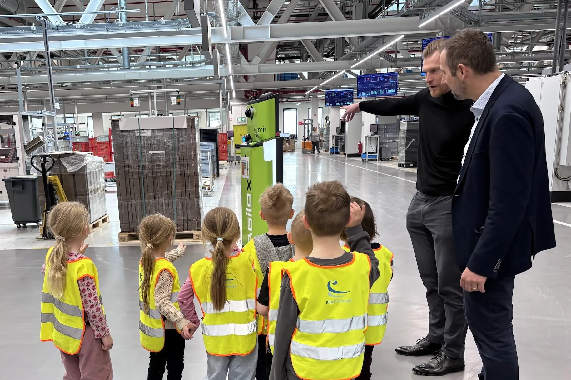 Ein Mann in Anzug und Krawatte erklärt einer Gruppe von Kindern in Warnwesten den Ablauf in einer großen Produktionshalle.