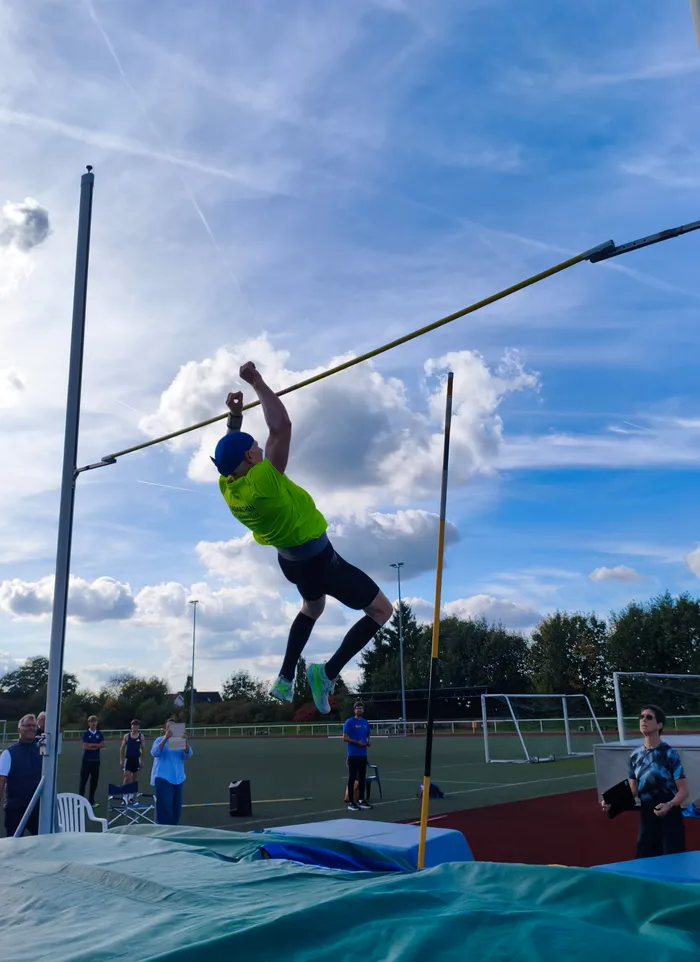 Ein Sportler in neongelbem T-Shirt und blauer Kappe springt über die Hochsprunglatte unter blauem Himmel.
