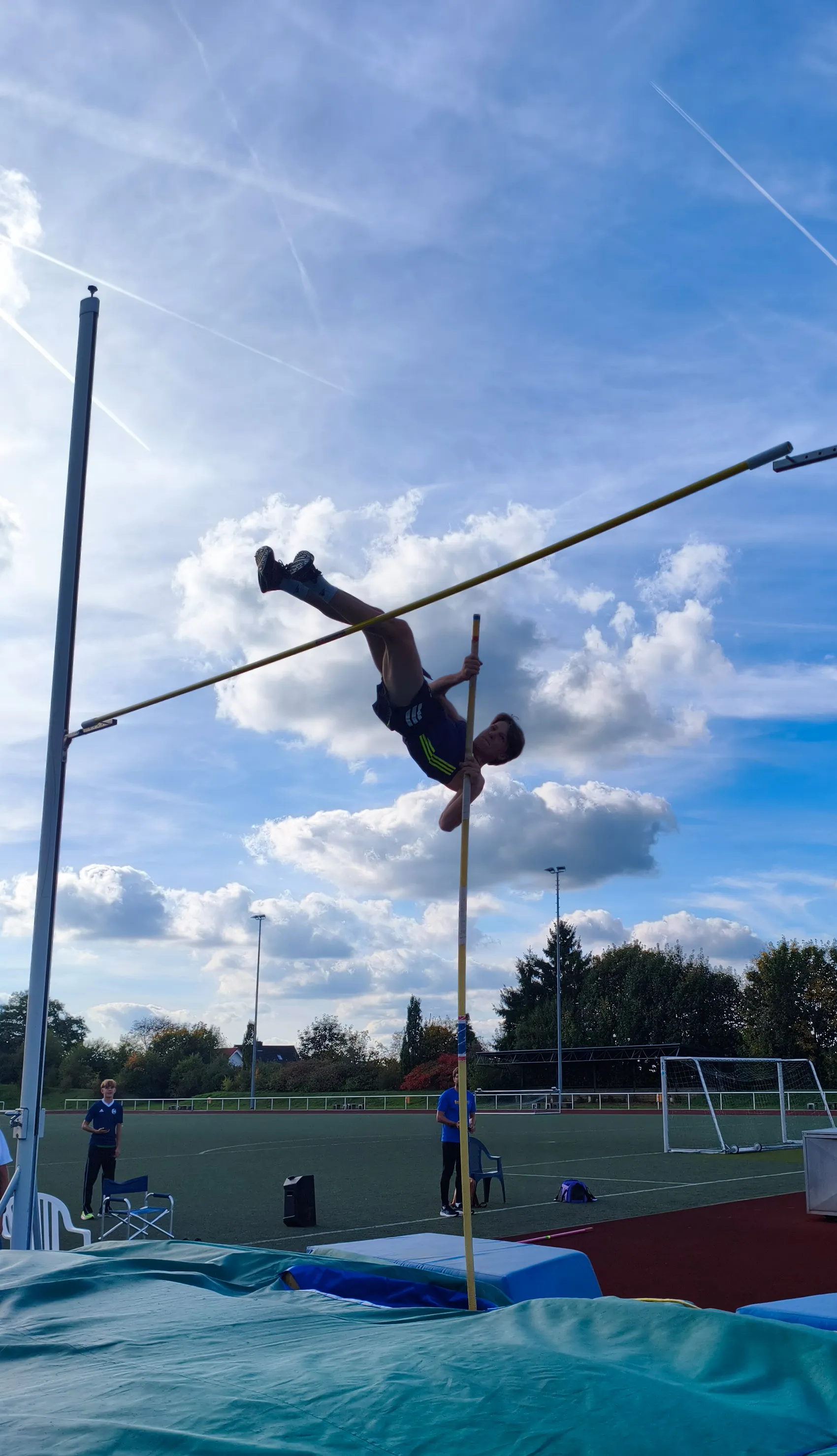 Ein Sportler überquert beim Stabhochsprung die Querlatte vor einem blau bewölkten Himmel.