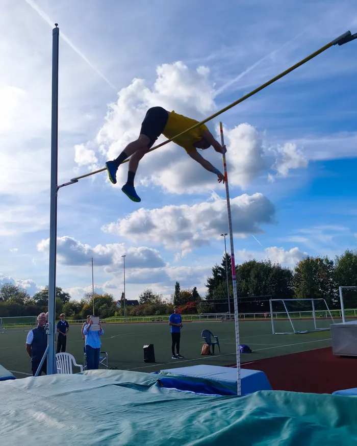 Ein Sportler in gelbem Trikot überquert beim Stabhochsprung die Latte auf einem Sportplatz unter blauem Himmel.