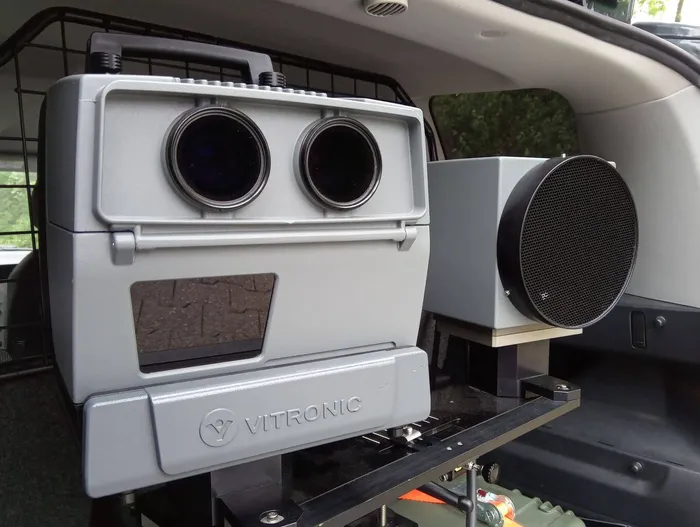 Ein graues Vitronic-Radargerät mit zwei Linsen und einem Display ist in einem Fahrzeug montiert.