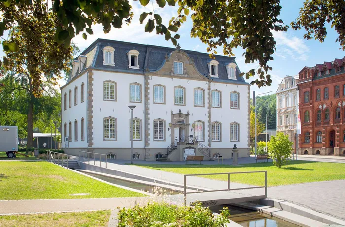 Das weiße Stadtmuseum Iserlohn mit dunklem Mansarddach vor einer Grünanlage und einem kleinen Wasserlauf.