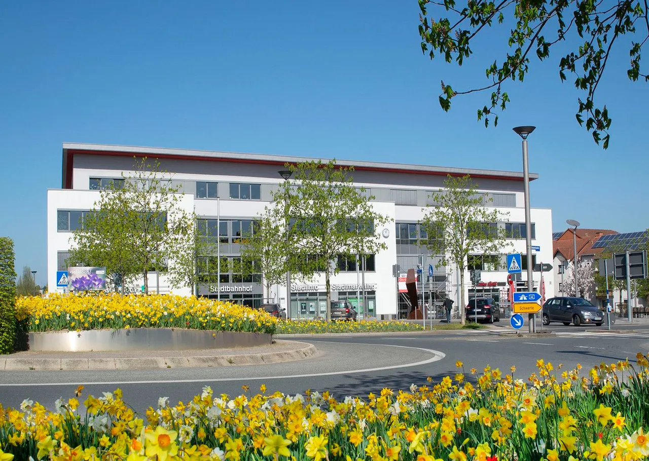 Ein weißes Gebäude des Stadtbahnhofs hinter einem Kreisverkehr mit gelben und weißen Narzissen unter blauem Himmel.