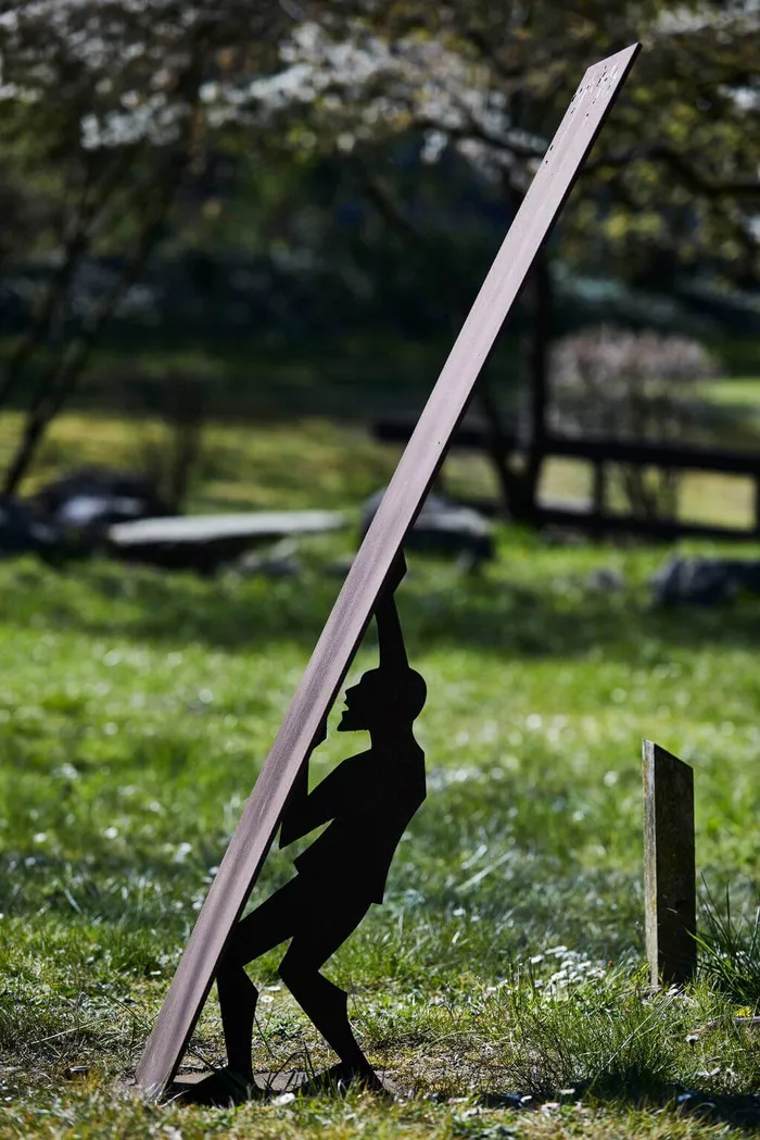 Eine Metallskulptur zeigt die Silhouette eines Mannes, der eine lange, schräg stehende Metallplatte stützt. Die Skulptur steht auf einer Rasenfläche im Freien.