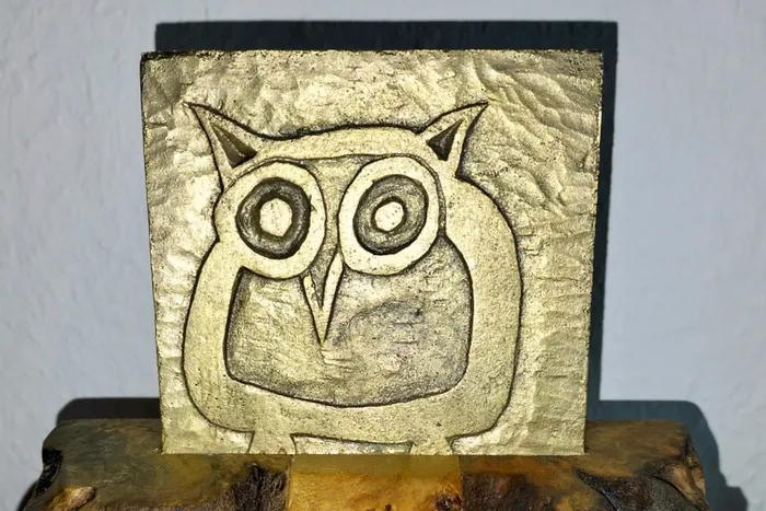 Eine quadratische, goldfarbene Plakette mit einer eingravierten Eule auf einem Holzsockel.