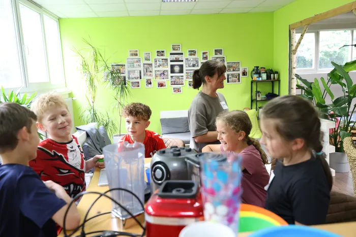 Kinder sitzen um einen Tisch mit einem Mixer und bereiten gesunde Snacks zu, während eine Betreuerin im Hintergrund lächelt. An der hellgrünen Wand hängen zahlreiche Fotos.