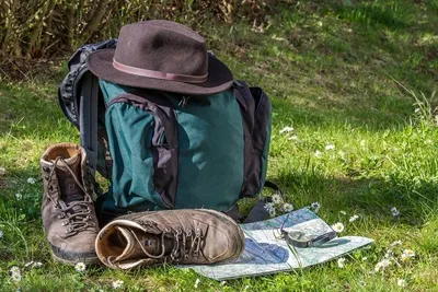 Ein grüner Rucksack mit braunem Hut, Wanderschuhe und eine Landkarte liegen auf einer Wiese.