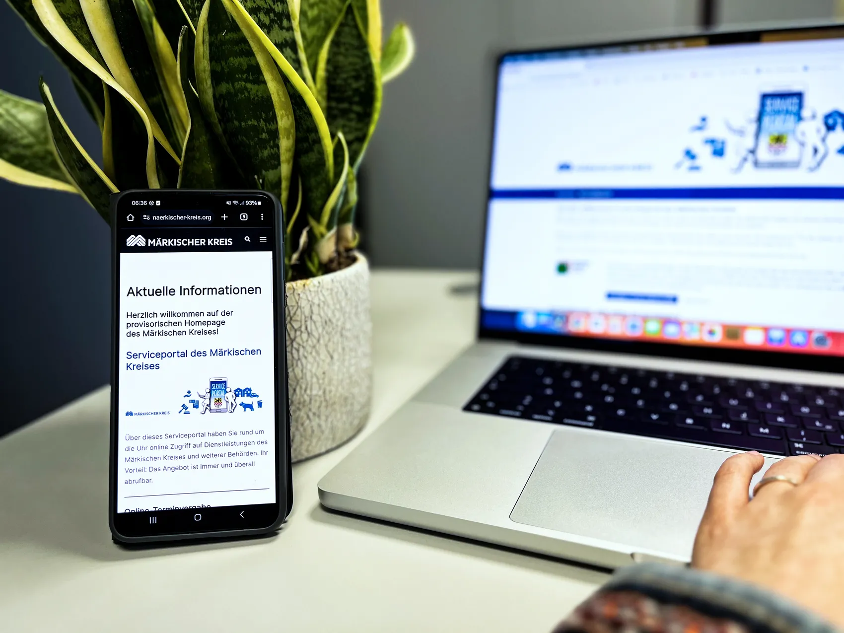 Smartphone zeigt die Webseite des Märkischen Kreises mit Informationen zum Serviceportal, daneben ein geöffnetes Laptop. Im Hintergrund eine Zimmerpflanze und ein Bild mit Menschen.