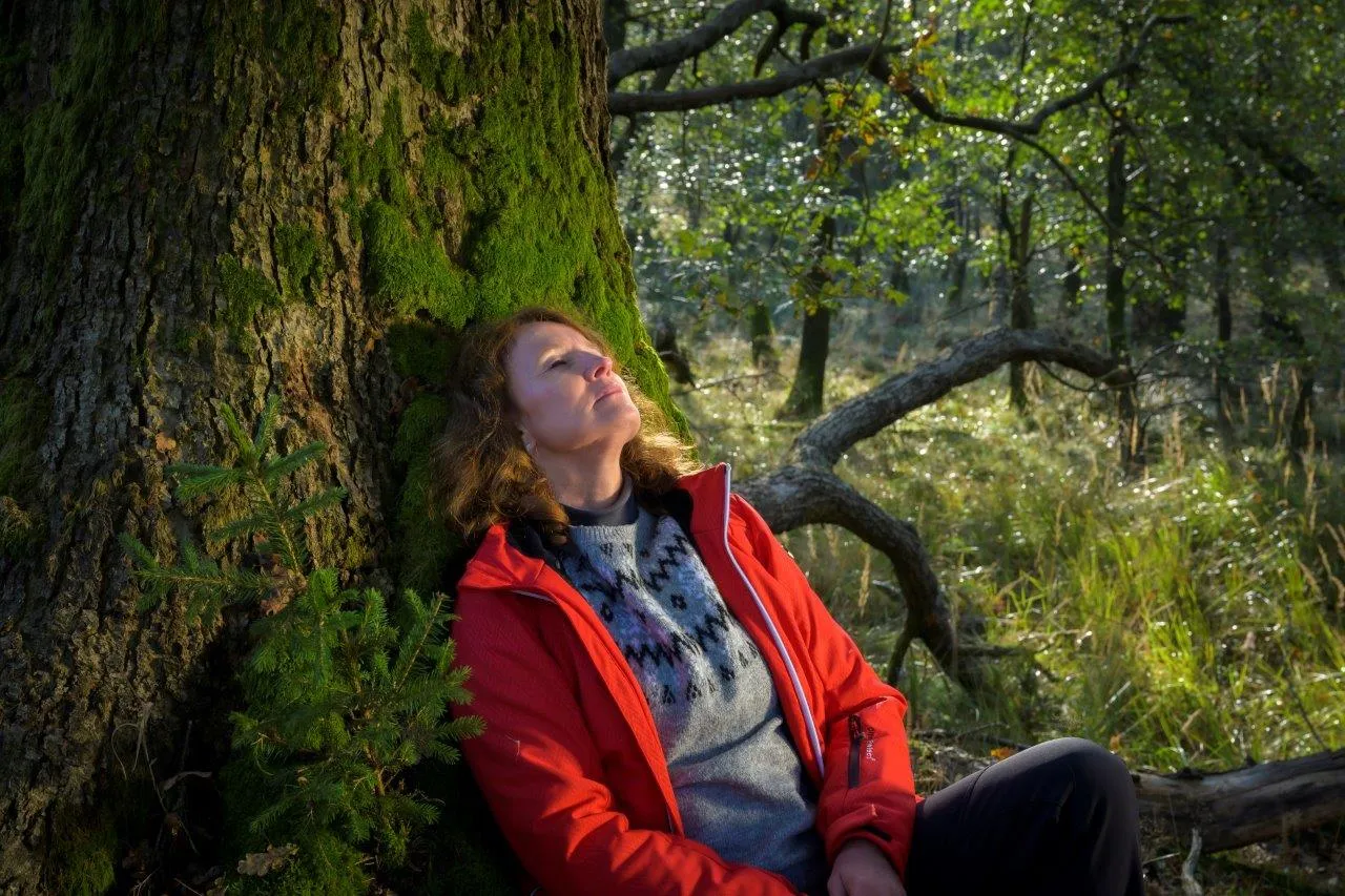 Eine Frau lehnt mit geschlossenen Augen an einem moosbewachsenen Baumstamm in einem Wald. Sie trägt eine rote Jacke und einen grauen Pullover.