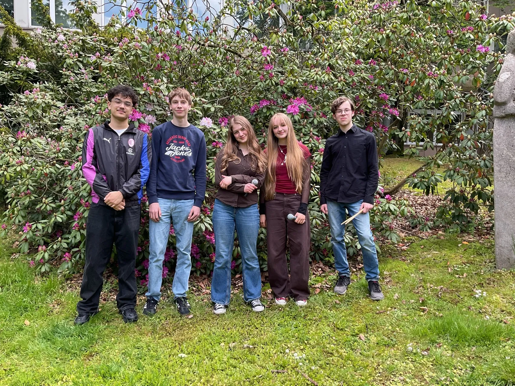 Fünf junge Musiker stehen auf einer grünen Wiese vor blühenden Büschen. Sie tragen Alltagskleidung und halten Instrumente wie Gitarren und einen Schlagstock.