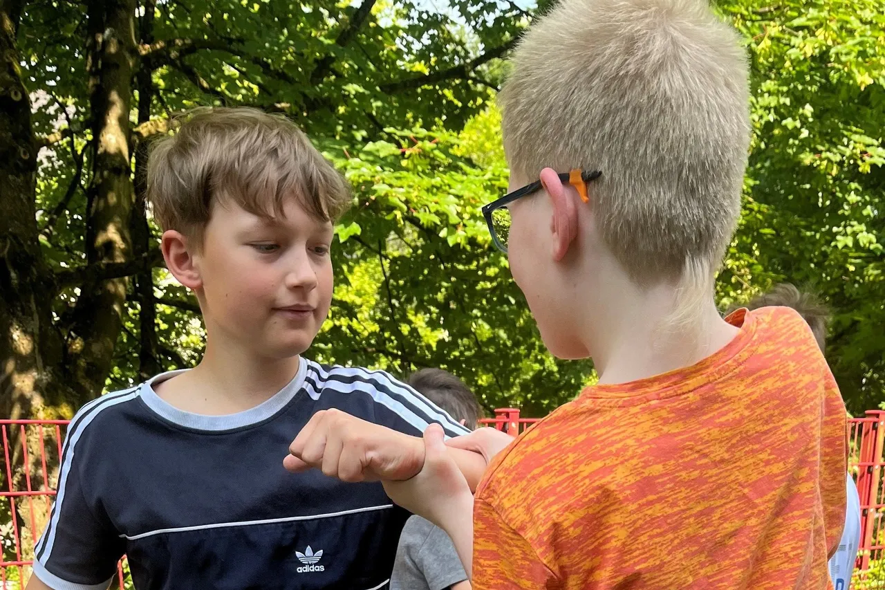 Zwei Jungen im Gespräch, einer zeigt dem anderen etwas auf seiner Hand. Im Hintergrund sind Bäume und ein roter Zaun zu sehen.