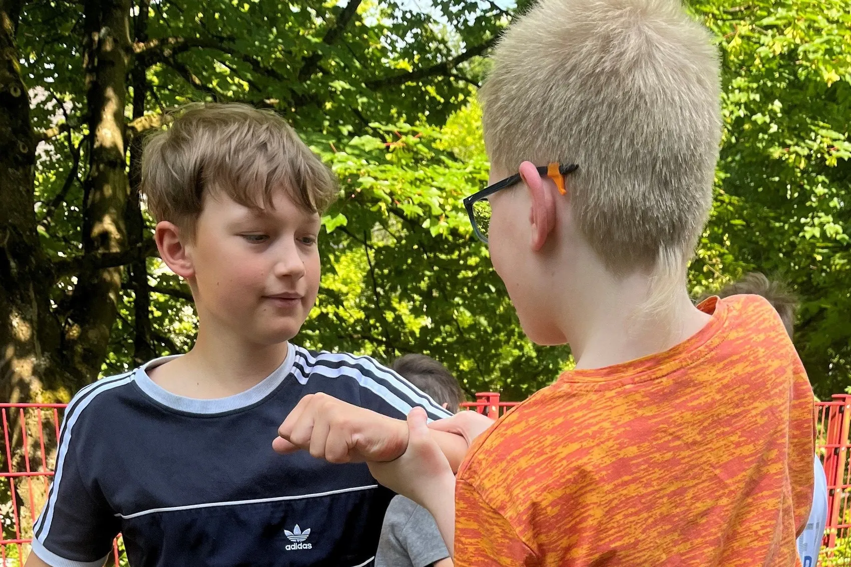 Zwei Jungen im Gespräch, einer zeigt dem anderen etwas auf seiner Hand. Im Hintergrund sind Bäume und ein roter Zaun zu sehen.