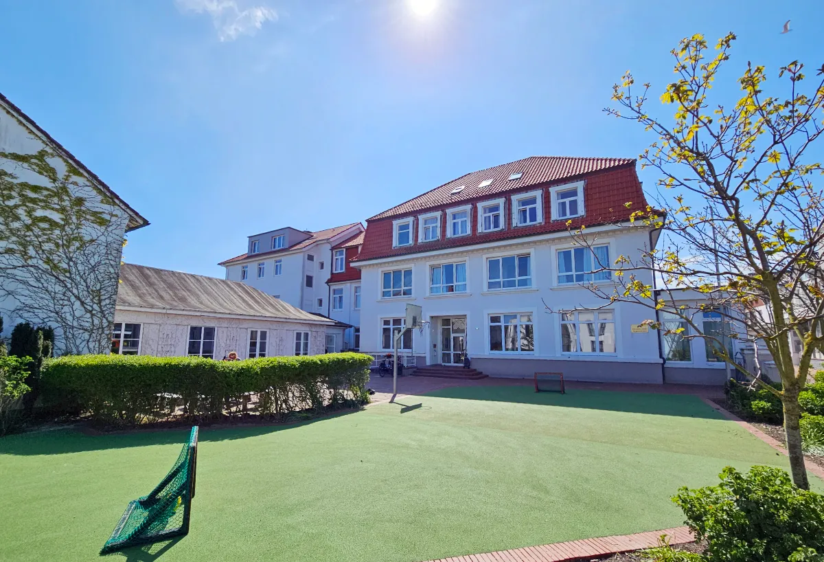 Das Schullandheim des Märkischen Kreises auf Norderney zeigt ein weißes Gebäude mit rotem Dach und einem gepflegten Gartenbereich. Im Vordergrund ist ein grüner Spielplatz mit einem kleinen Fußballtor zu sehen.