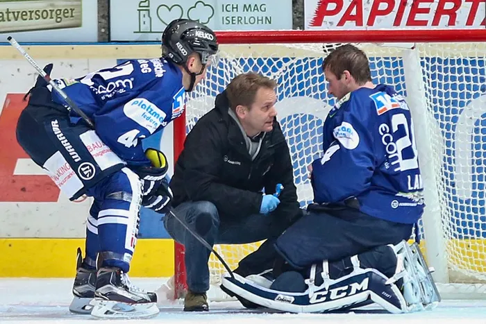 Mannschaftsarzt Dr. Jochen Veit kniet auf dem Eis und untersucht den sitzenden Torwart Mathias Lange.
