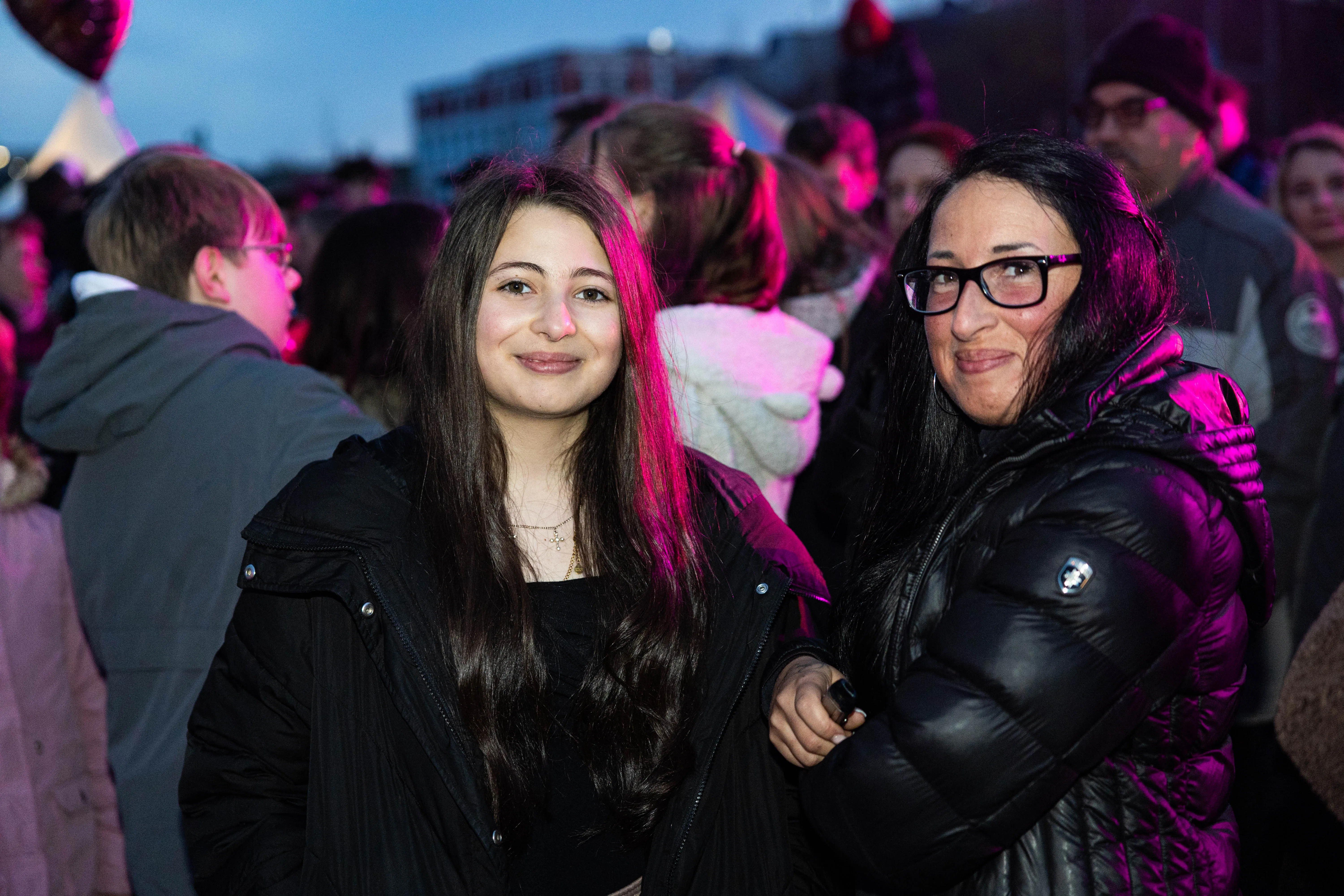 Zwei Frauen lächeln in die Kamera, umgeben von einer Menschenmenge bei einer Veranstaltung mit pinkfarbenem Licht. Im Hintergrund sind Gebäude und weitere Besucher zu erkennen.