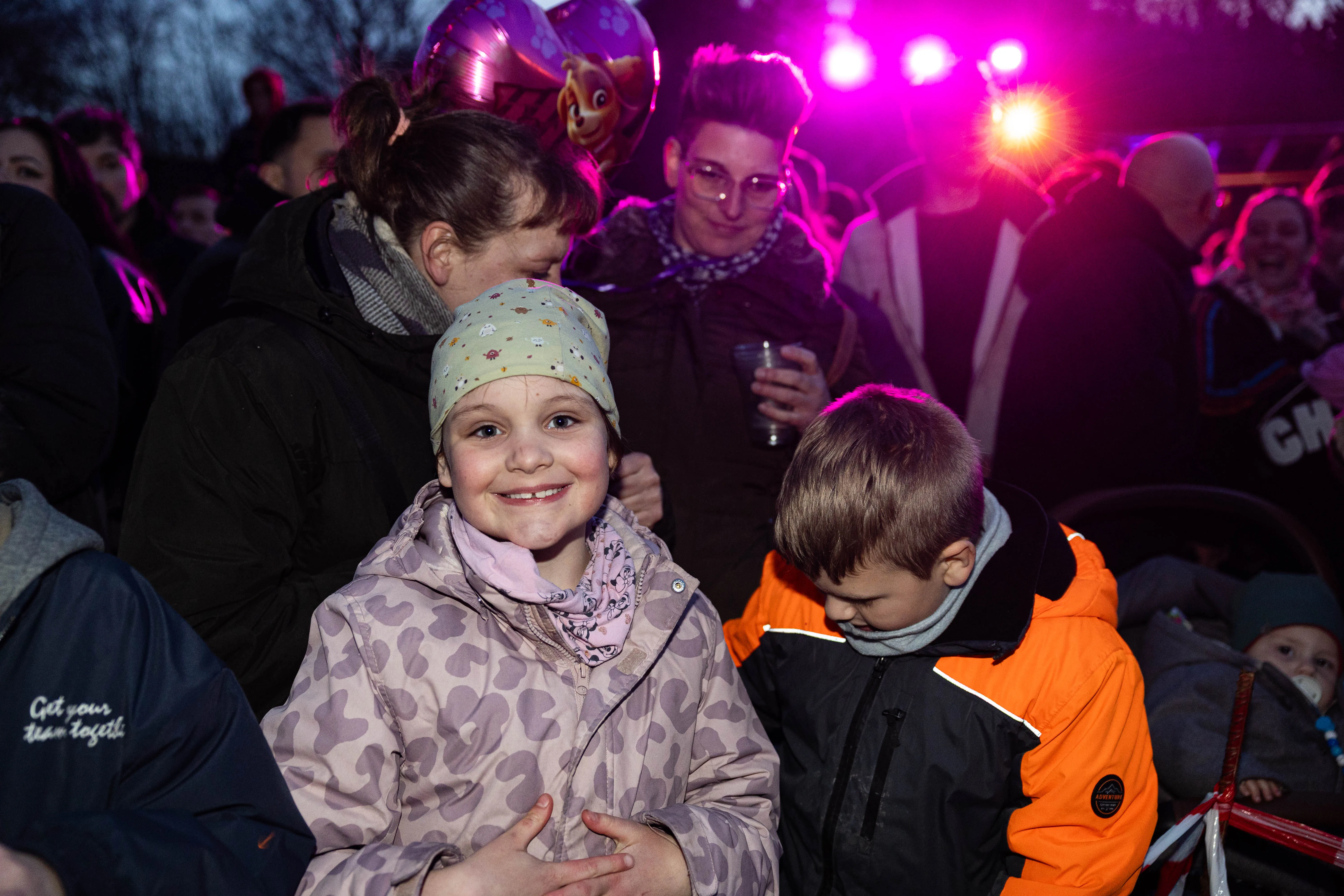 Zwei Kinder lächeln in die Kamera, umgeben von Menschenmenge und Lichtern auf einem Osterfeuer. Im Hintergrund sind weitere Besucher und ein roter Kinderwagen zu sehen.