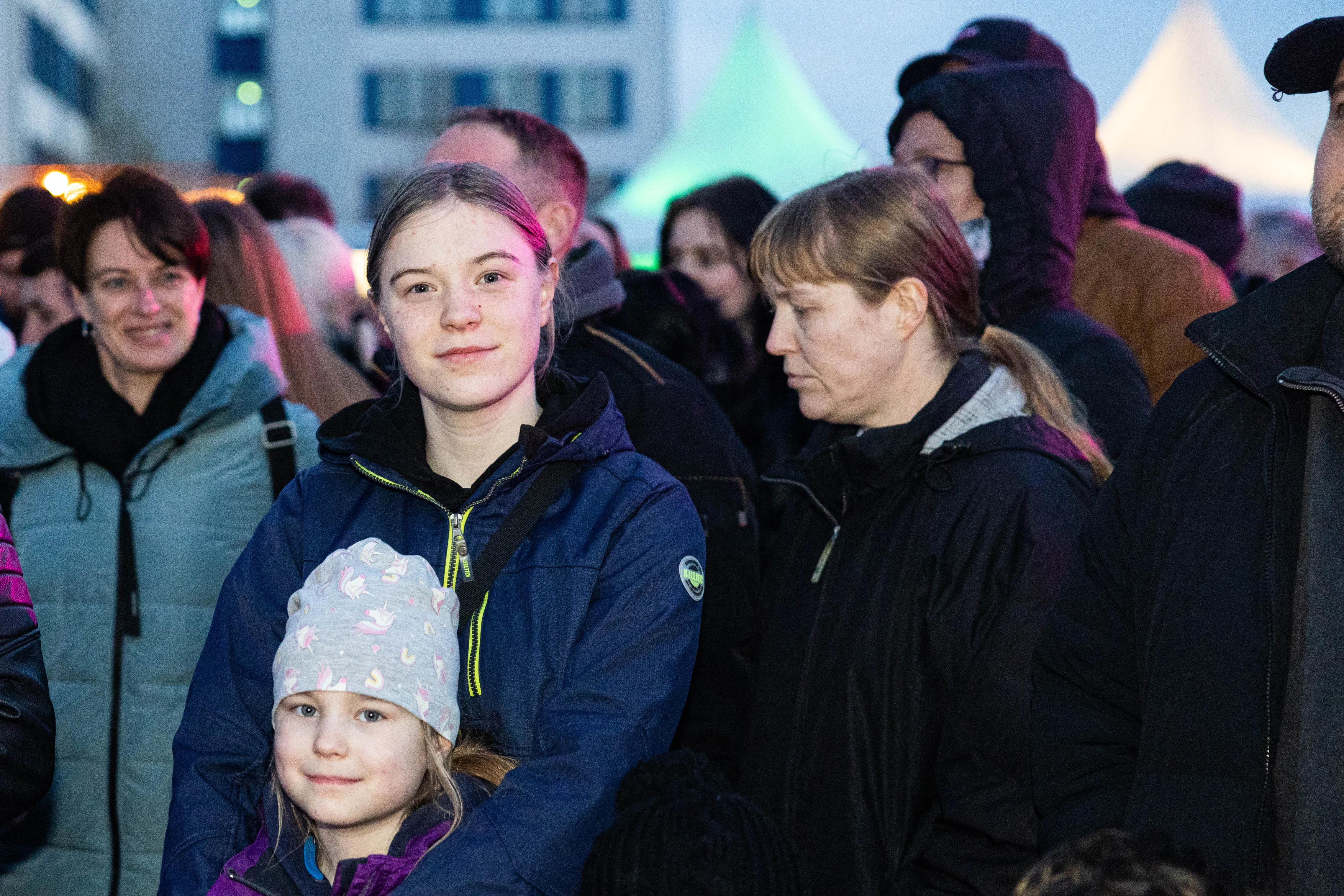 Drei Frauen und ein Mädchen stehen vor einer Menschenmenge, im Hintergrund ein helles Licht. Sie blicken in unterschiedliche Richtungen.