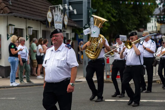 Stefan Schneider in weißem Hemd und schwarzer Baskenmütze vor einer marschierenden Blaskapelle mit Blechblasinstrumenten.