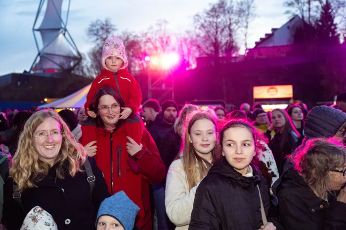Menschenmenge vor einem Osterfeuer, eine Frau trägt ein Kind auf ihren Schultern. Im Hintergrund ist ein großes, dreieckiges Gerüst zu sehen.