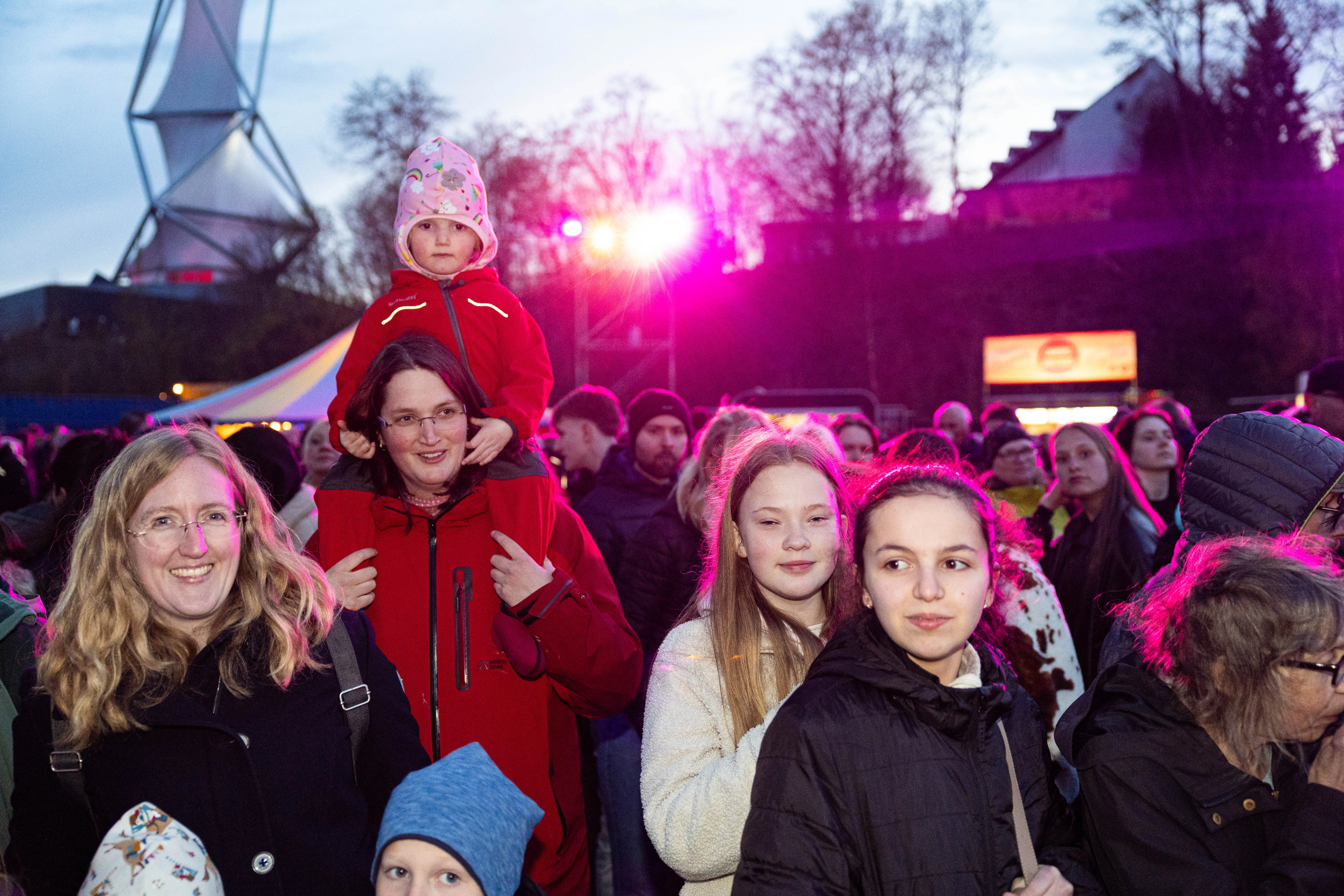 Menschenmenge vor einem Osterfeuer, eine Frau trägt ein Kind auf ihren Schultern. Im Hintergrund ist ein großes, dreieckiges Gerüst zu sehen.