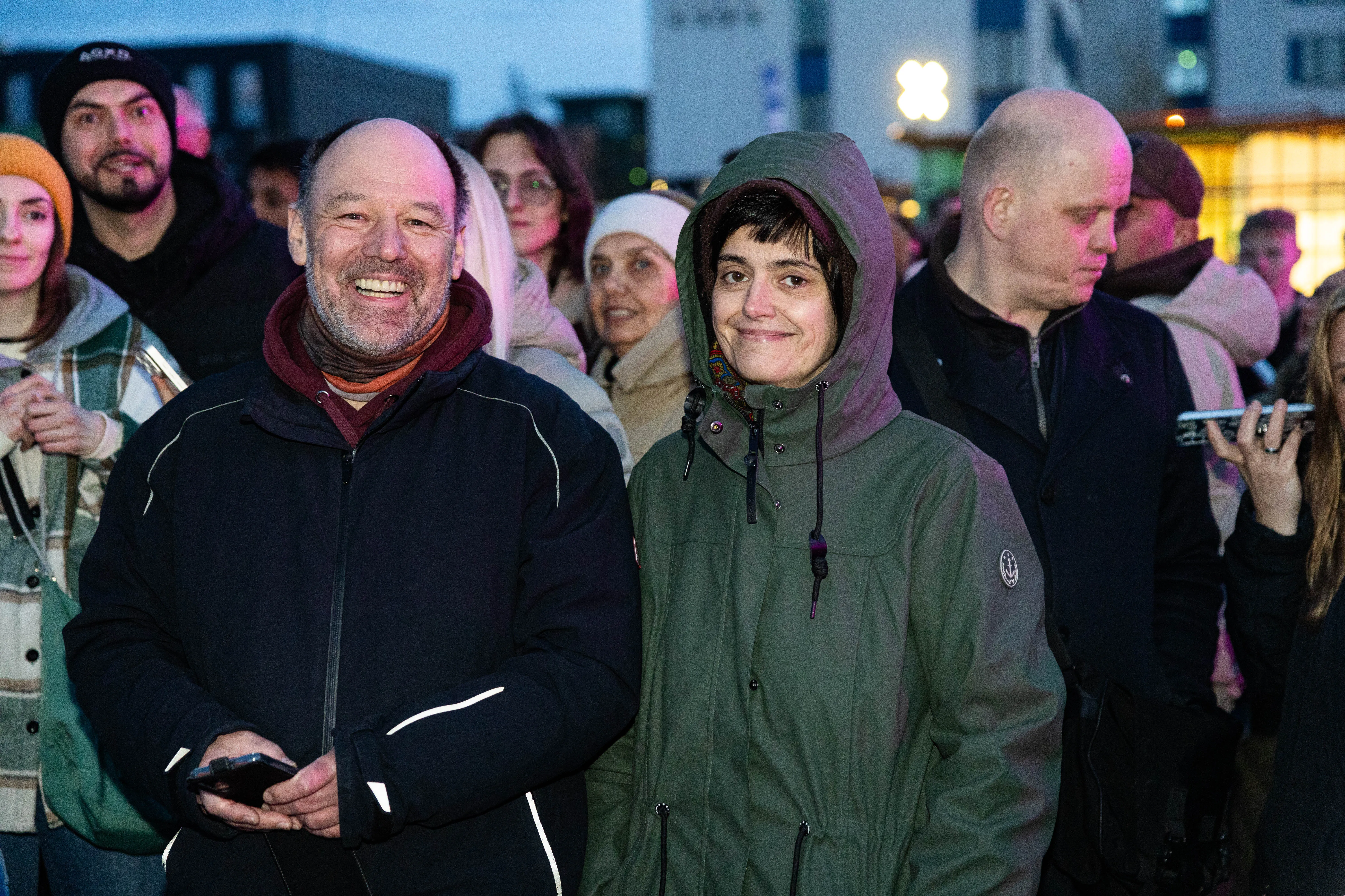 Zwei lächelnde Menschen im Vordergrund, ein Mann mit Glatze und ein rotem Halstuch sowie eine junge Frau mit grüner Jacke und Kapuze, umgeben von einer Menschenmenge bei einer Veranstaltung im Freien.