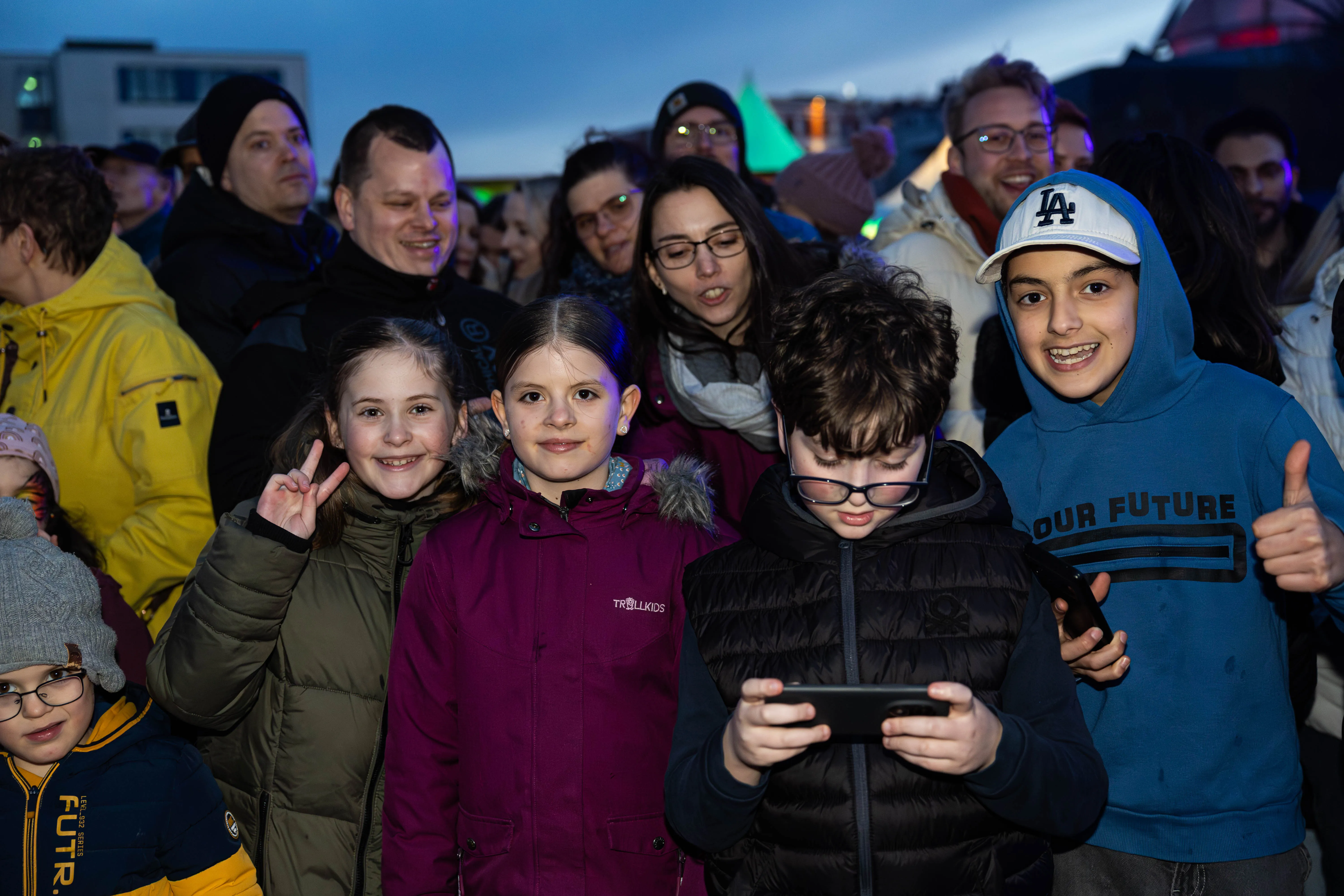 Mehrere Kinder und Erwachsene schauen gemeinsam auf ein Smartphone, während im Hintergrund ein Osterfeuer stattfindet.
