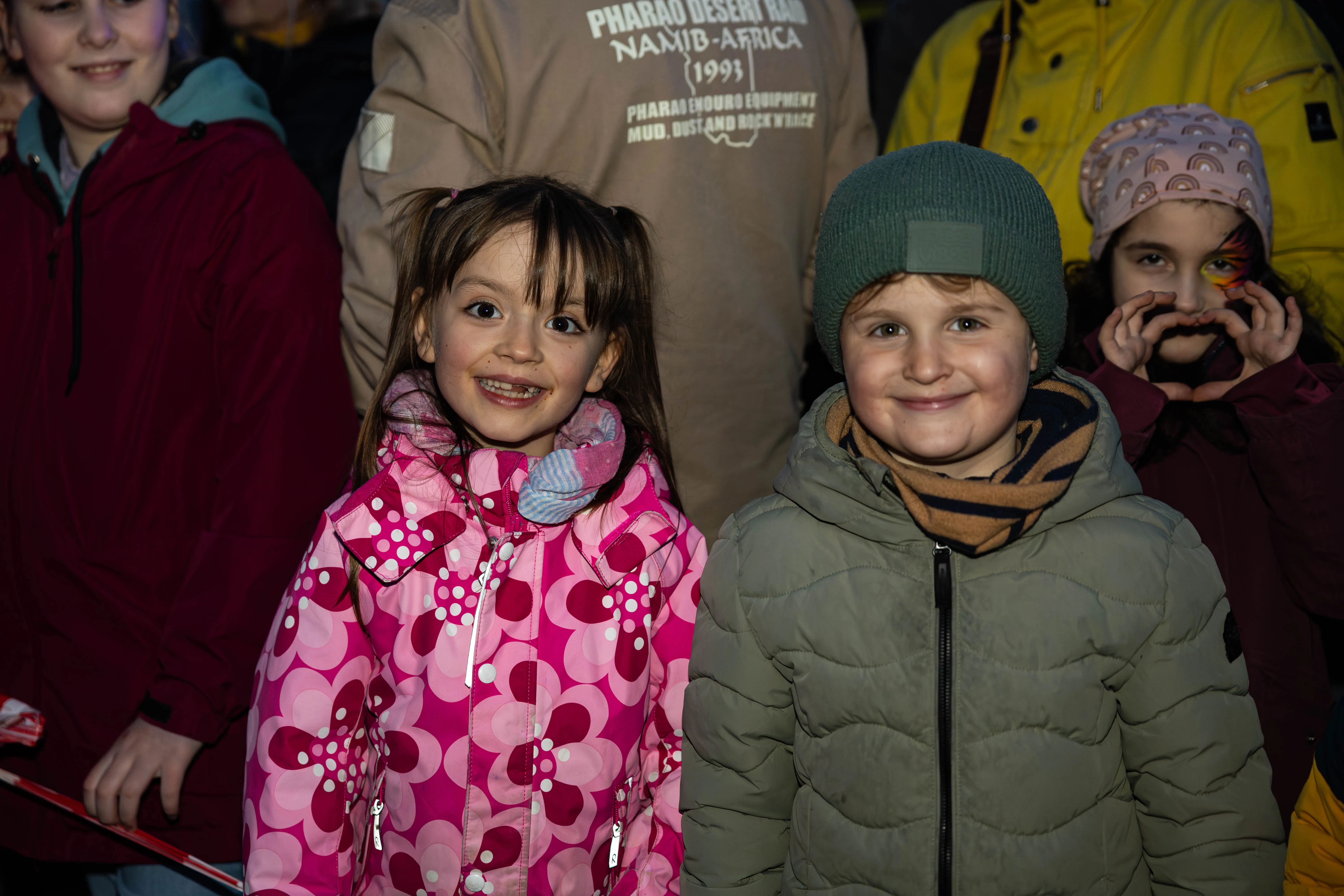Zwei lächelnde Kinder im Vordergrund, ein Mädchen in pinker Jacke und ein Junge in olivgrüner Jacke, umgeben von weiteren Menschen bei einer Veranstaltung.