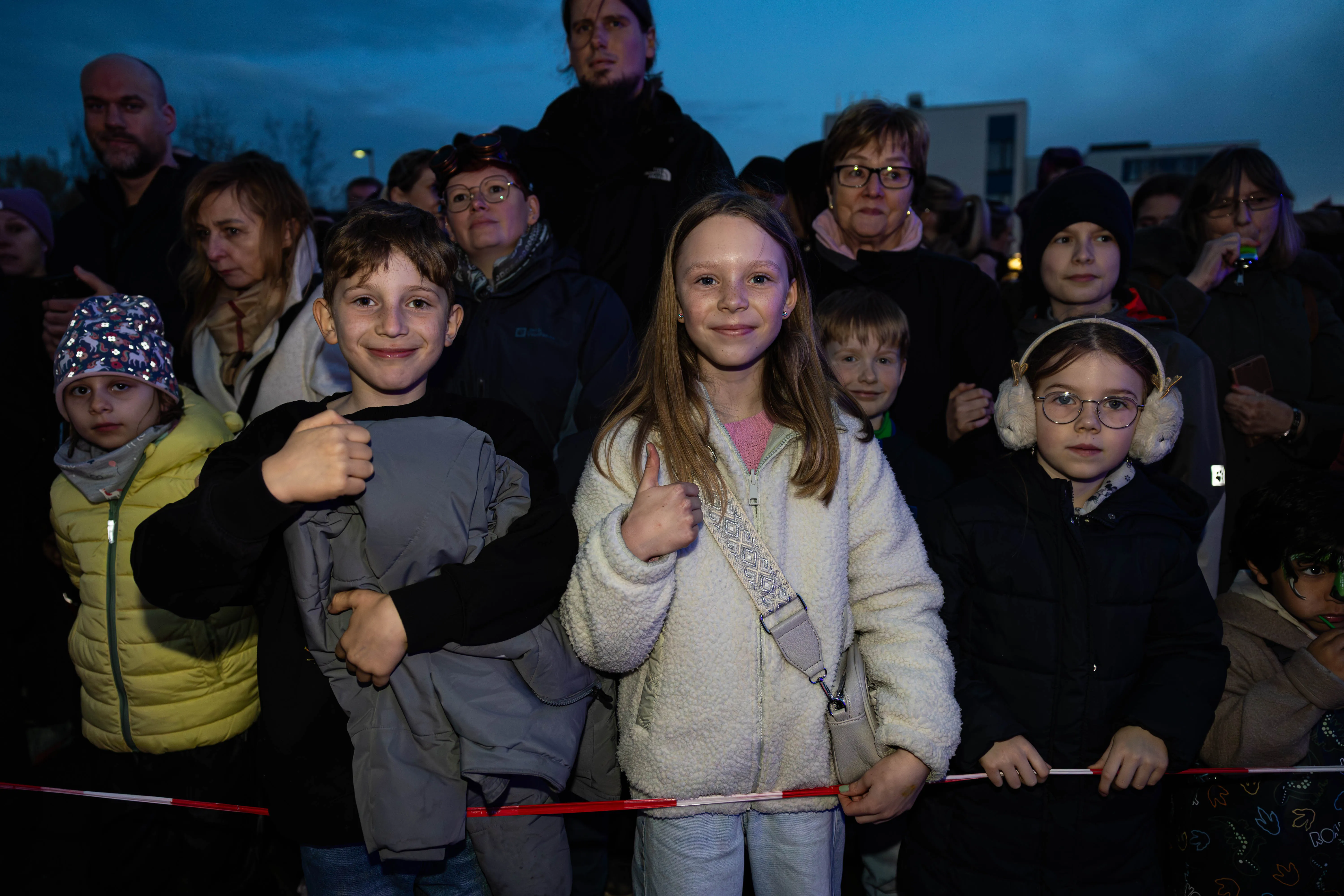 Mehrere Kinder und Erwachsene stehen vor einer Absperrung und schauen in die Ferne, einige zeigen Daumen hoch. Im Hintergrund sind weitere Menschen und Lichtquellen zu sehen.