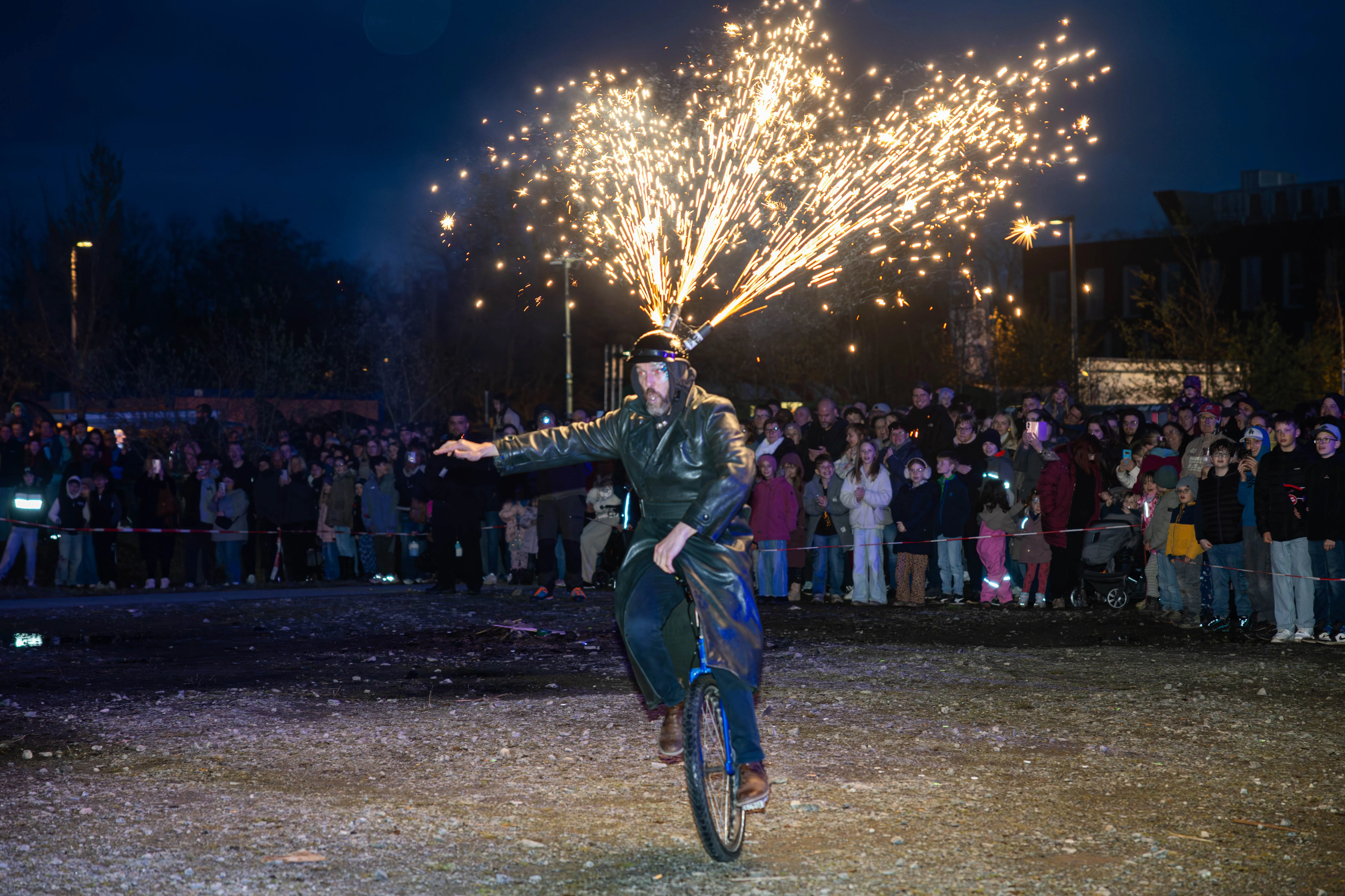 Ein Mann fährt auf einem Einrad, während aus einem Feuerwerk auf seinem Kopf Funken sprühen. Im Hintergrund steht eine große Menschenmenge und beobachtet die Darbietung.