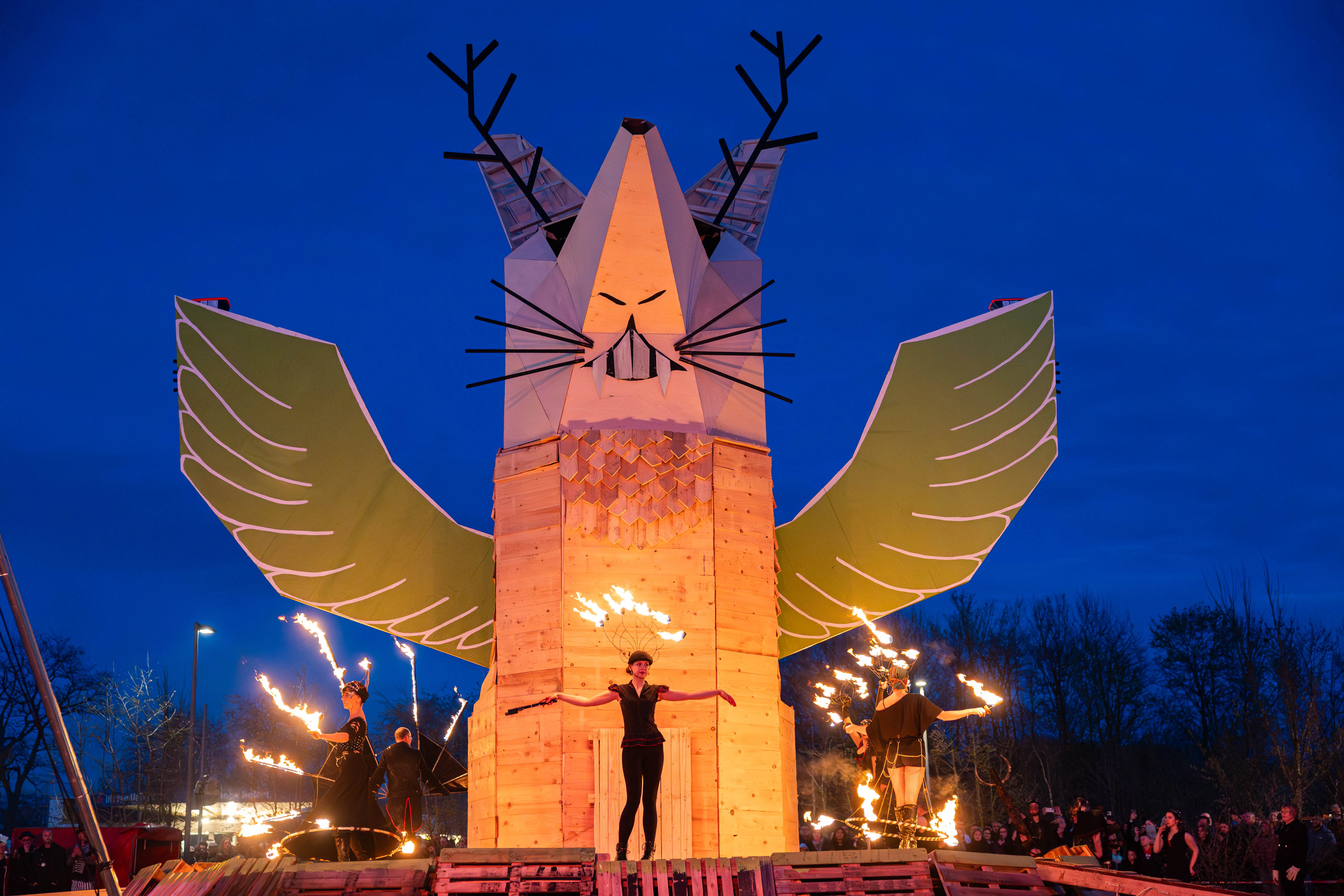 Große, leuchtende Holzskulptur eines Fuchses mit Geweih und Flügeln steht im Mittelpunkt eines Osterfeuers. Vor der Skulptur steht eine Person mit ausgebreiteten Armen, während andere Personen mit Feuer jonglieren.