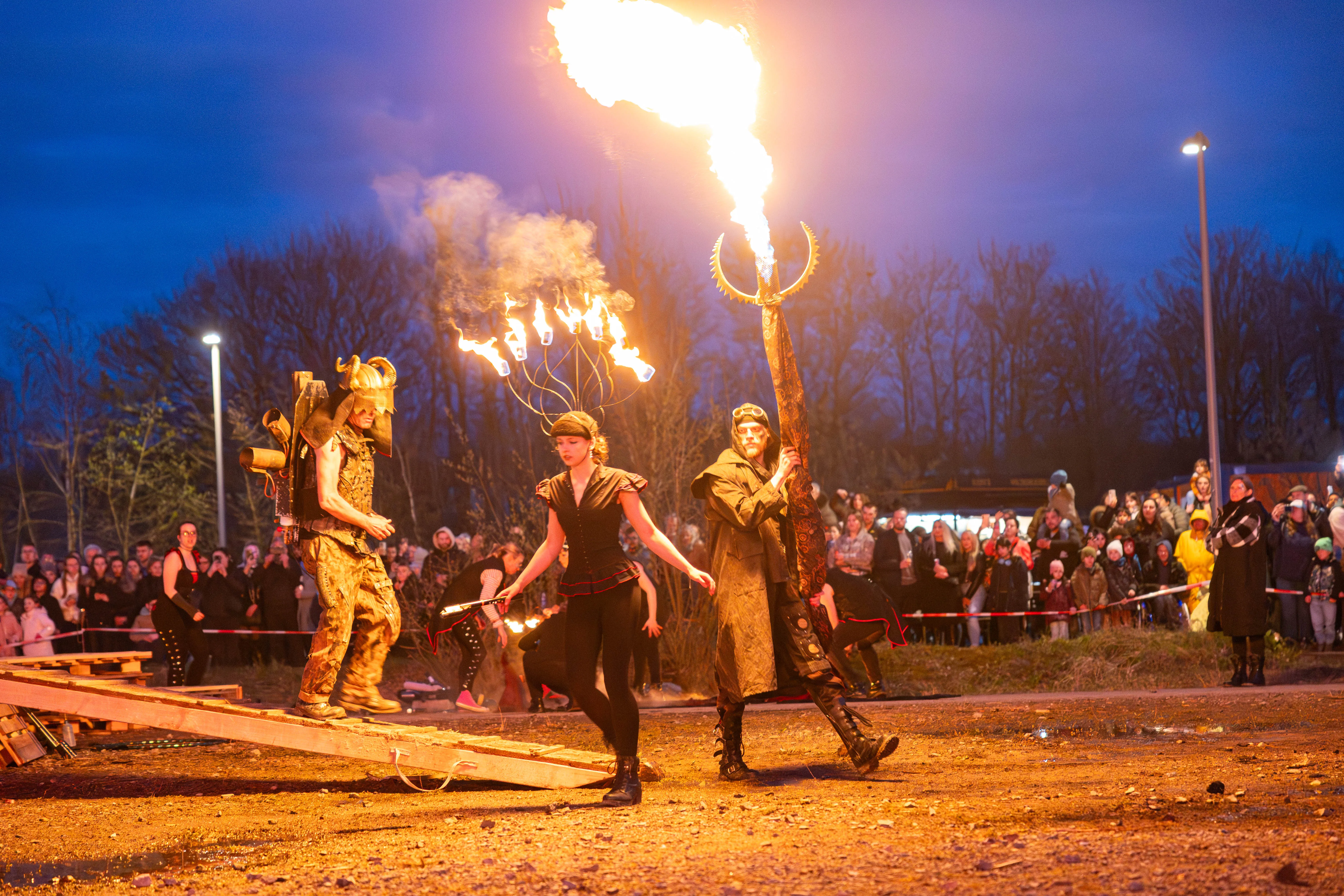 Drei Personen in Kostümen mit Feuer jonglieren vor einer großen Menschenmenge bei einer Osterfeuer-Veranstaltung in Lüdenscheid.