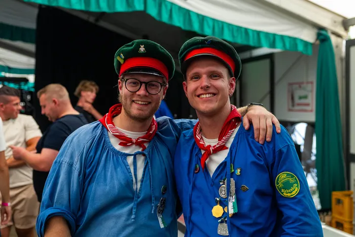 Zwei Männer der Meinerzhagener Schützengilde in blauen Hemden, roten Halstüchern und grünen Mützen lächeln in die Kamera.