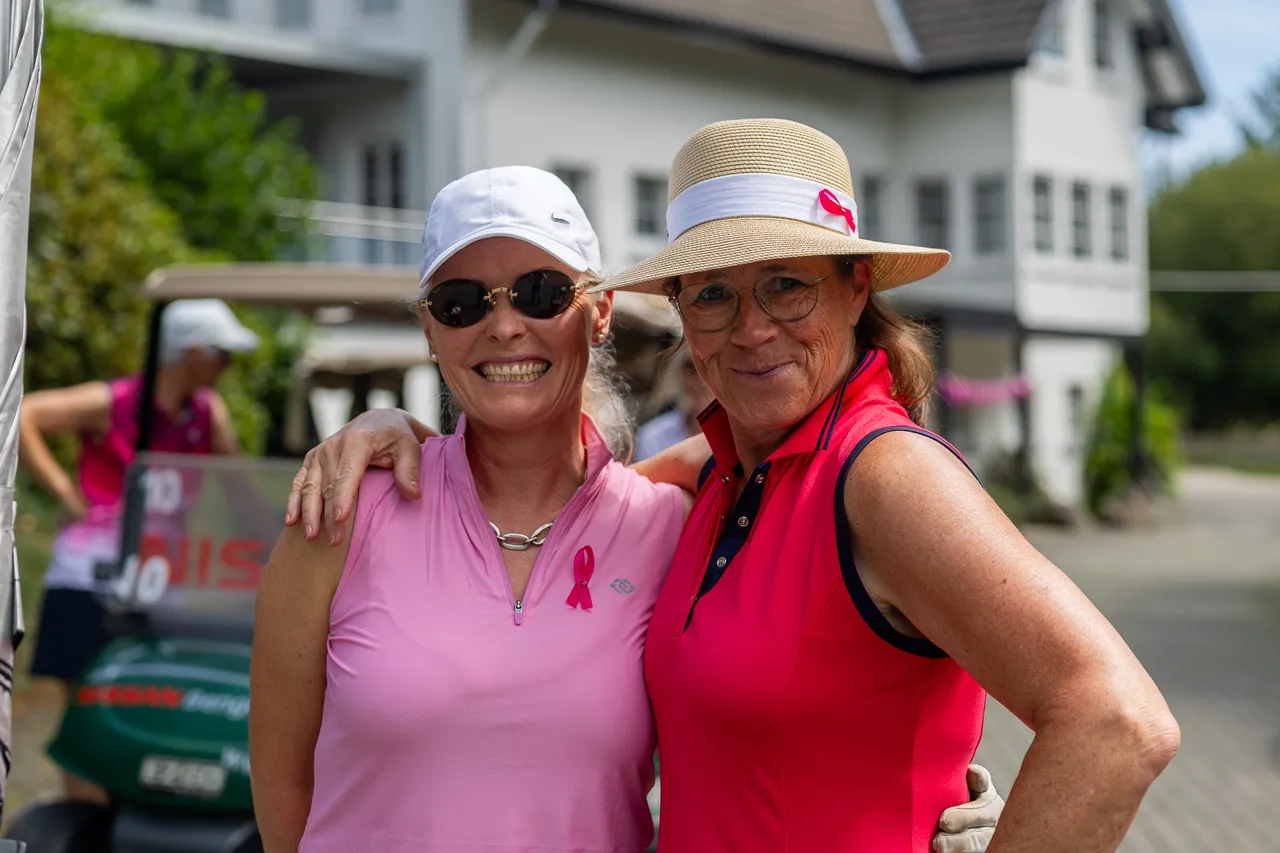 Zwei lächelnde Frauen in pinkfarbener Golfbekleidung posieren vor einem Gebäude und einem Golfcart.