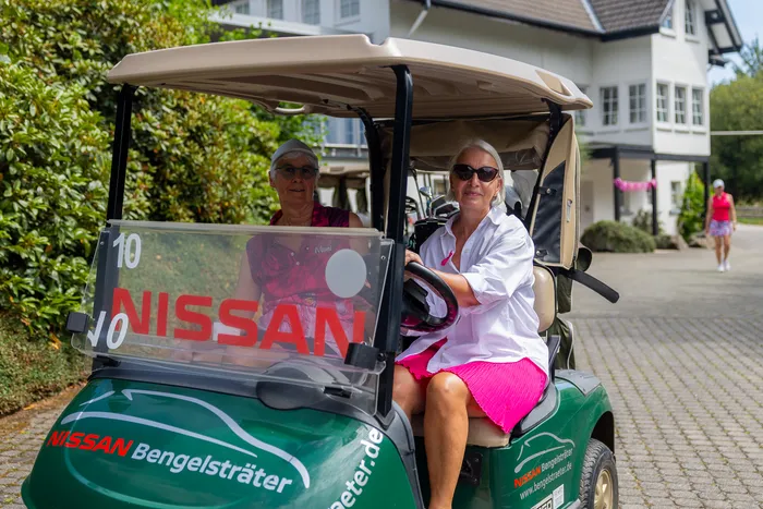 Zwei Frauen sitzen in einem grünen Golfkarren und tragen pinkfarbene Kleidung.