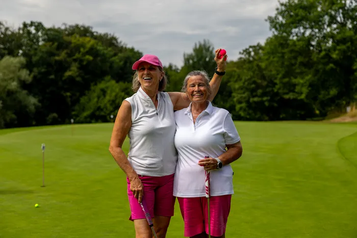 Zwei lächelnde Frauen in weißer Sportkleidung und pinkfarbenen Hosen stehen auf einem Golfplatz; eine hält einen pinkfarbenen Golfball in die Höhe.