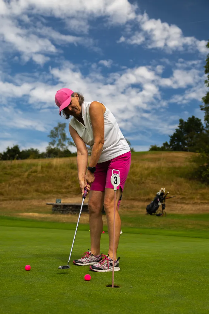 Eine Frau in weißem Oberteil und pinker Sportkleidung puttet einen Golfball auf einem Golfplatz.