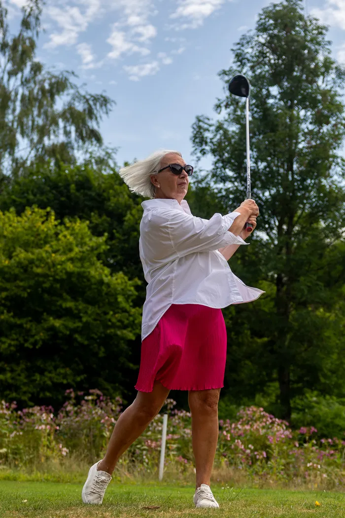 Eine Frau mit weißen Haaren schwingt einen Golfschläger auf einem Rasenplatz. Sie trägt ein weißes Hemd und einen pinkfarbenen Rock.