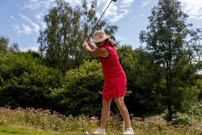 Eine Frau in einem roten Kleid und einem hellen Hut schwingt einen Golfschläger auf einem Golfplatz.