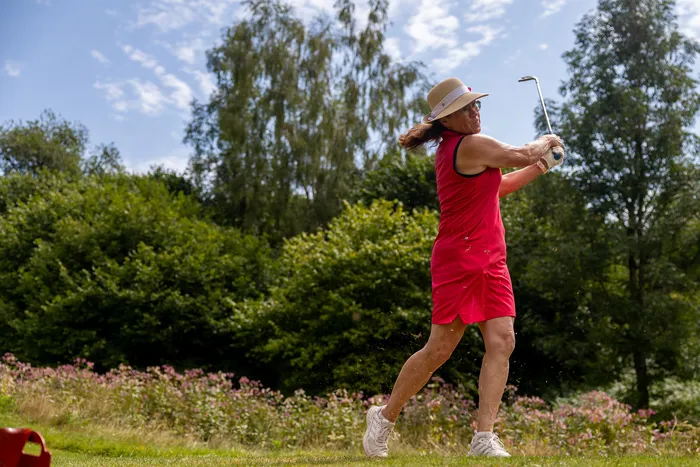 Eine Golferin in einem roten Kleid und Sonnenhut schwingt einen Golfschläger auf einer Wiese vor Bäumen und Blumen.