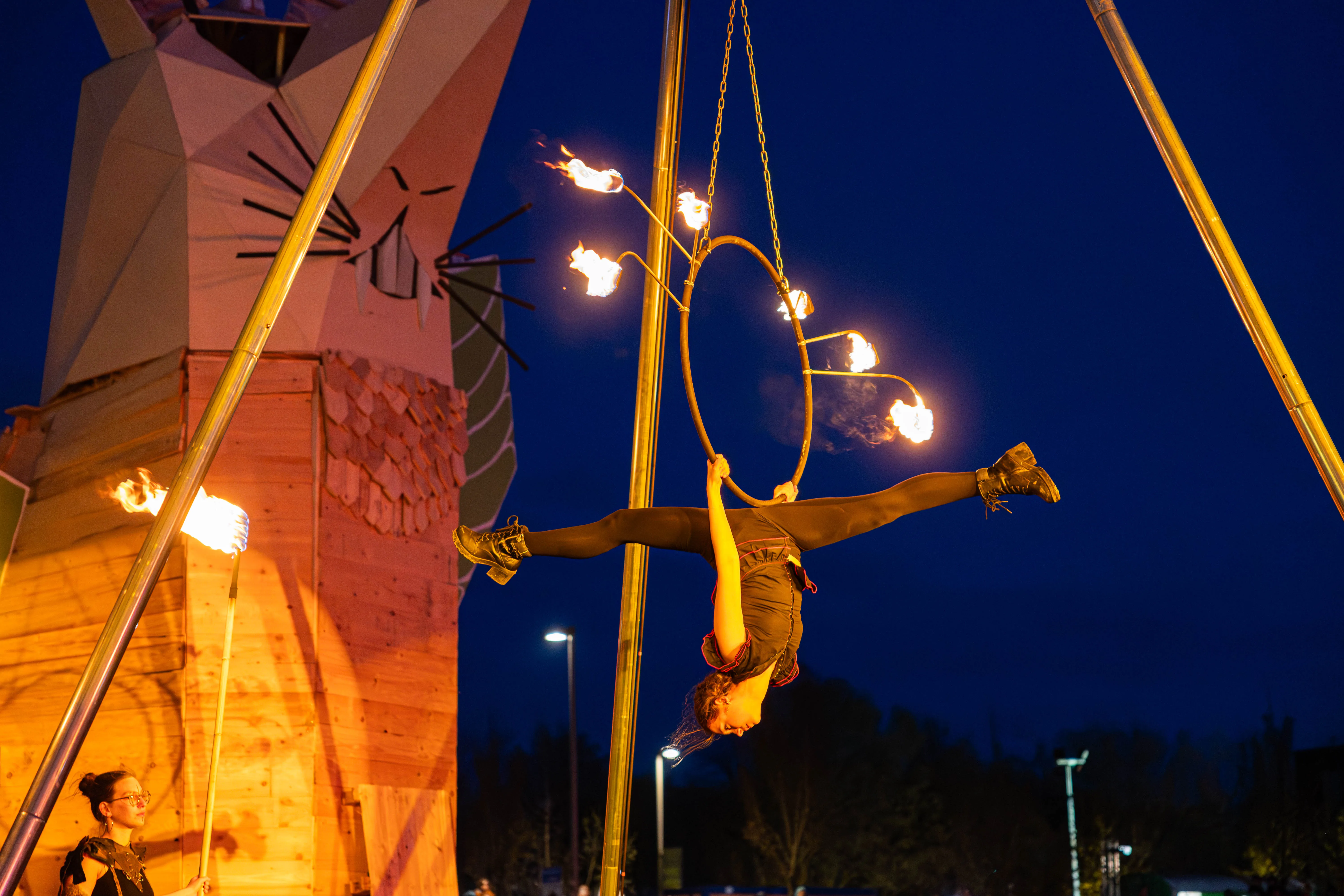 Eine Akrobatin hängt an einem brennenden Reifen, der an einer Metallkonstruktion aufgehängt ist. Im Hintergrund ein großes Holzgebäude in Katzenform und ein dunkler Nachthimmel.